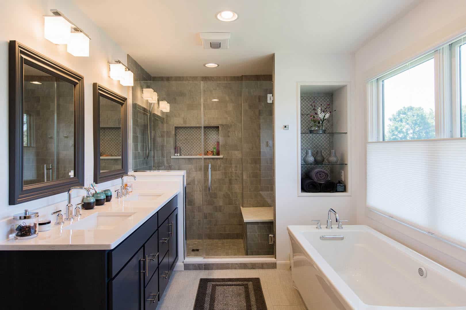 Modern bathroom with double sinks, a large mirror, dark cabinets, a glass-enclosed shower with a bench, and a white bathtub near a window with blinds. Decorative shelves hold towels and accessories.