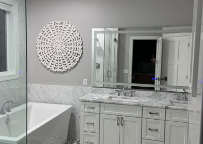 Modern bathroom with a white bathtub, marble accents, patterned tile floor, white double-sink vanity, large mirror, decorative wall art, and a partially open door—showcasing how collaborating with homeowners leads to truly transforming homes.