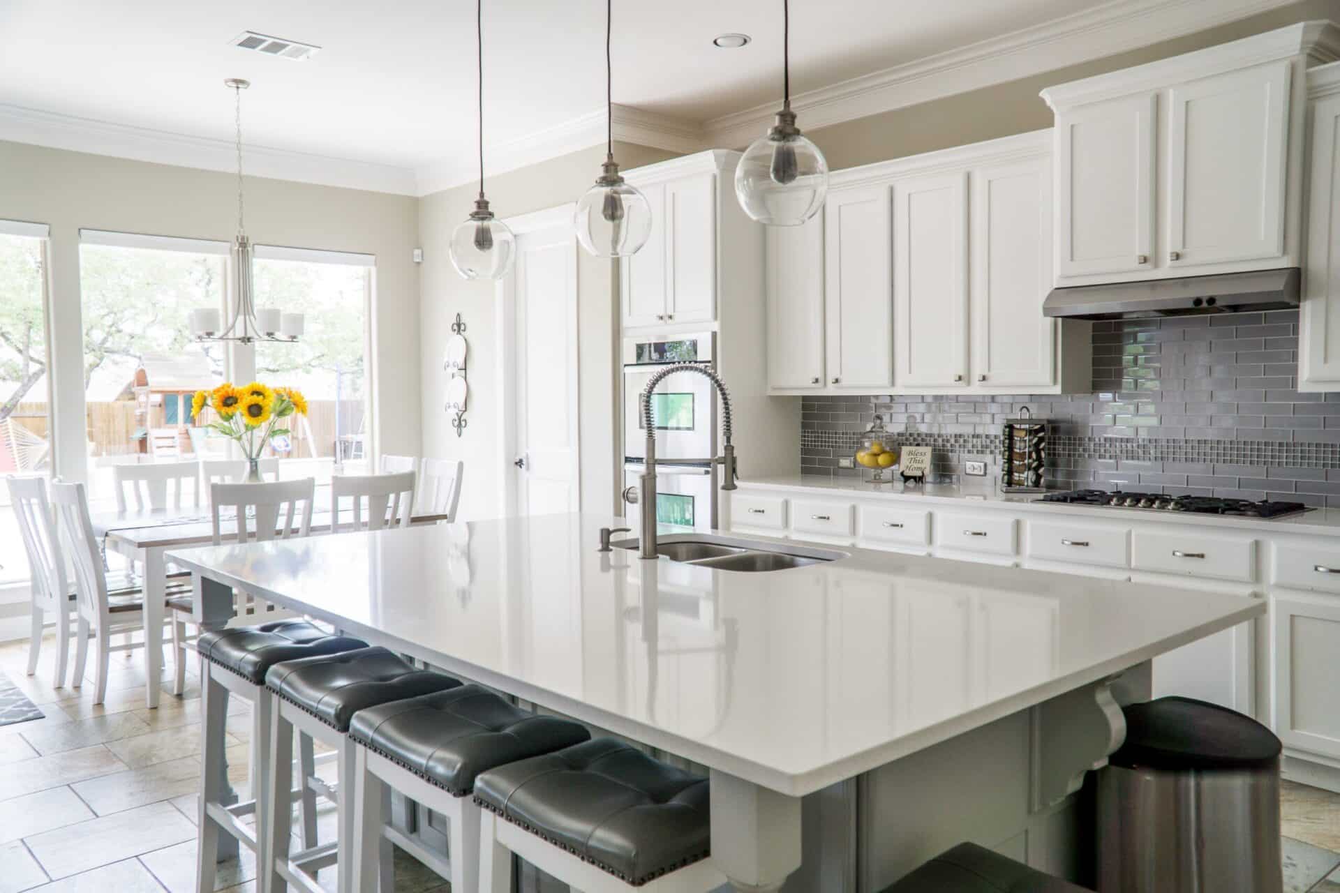 Bright modern kitchen with white cabinets, a large white island with gray cushioned stools, stainless steel appliances, pendant lights, and a dining area with a table and vase of sunflowers by large windows.