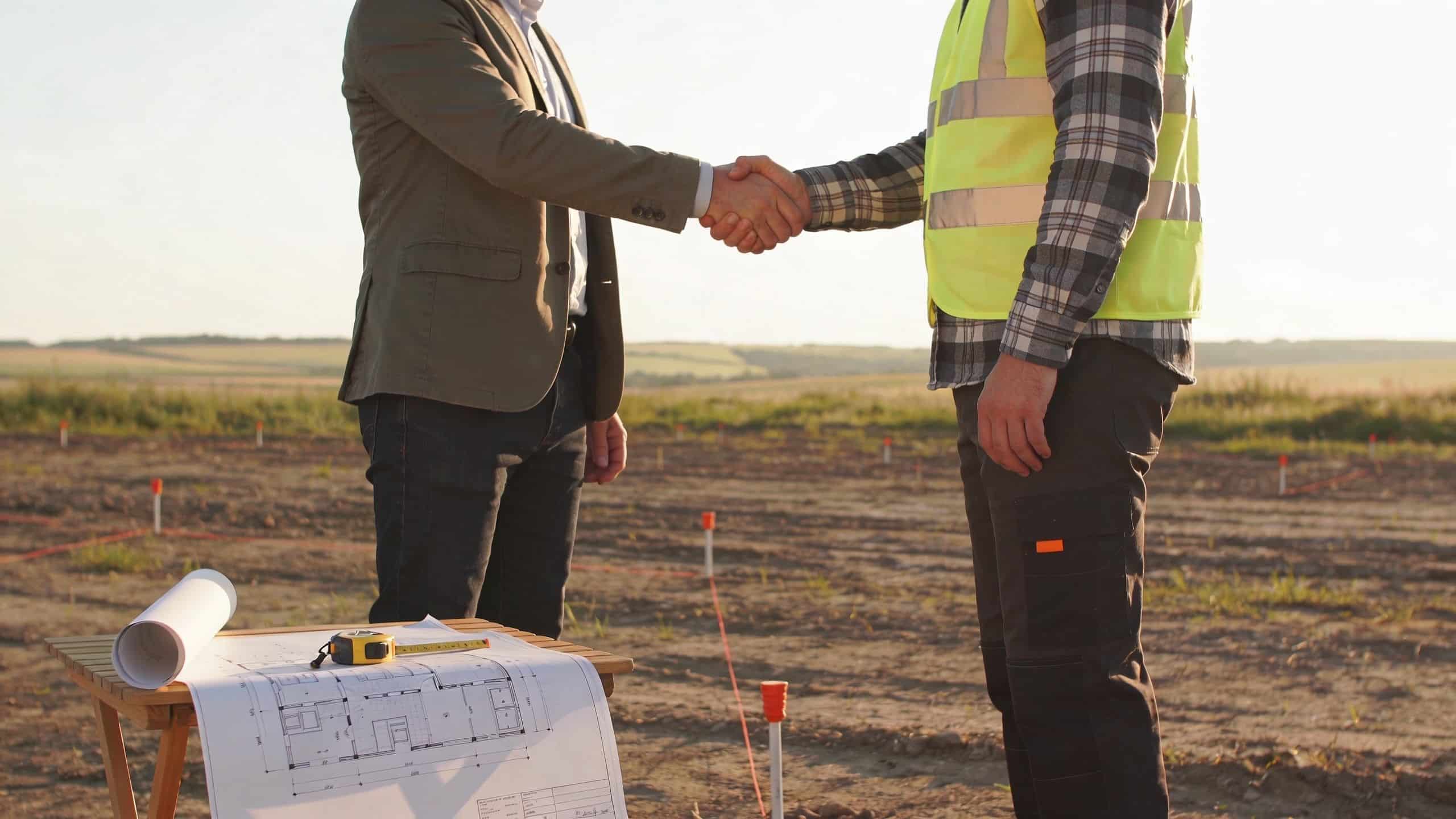 A businessman in a suit shakes hands with a construction worker at a building site with blueprints.
