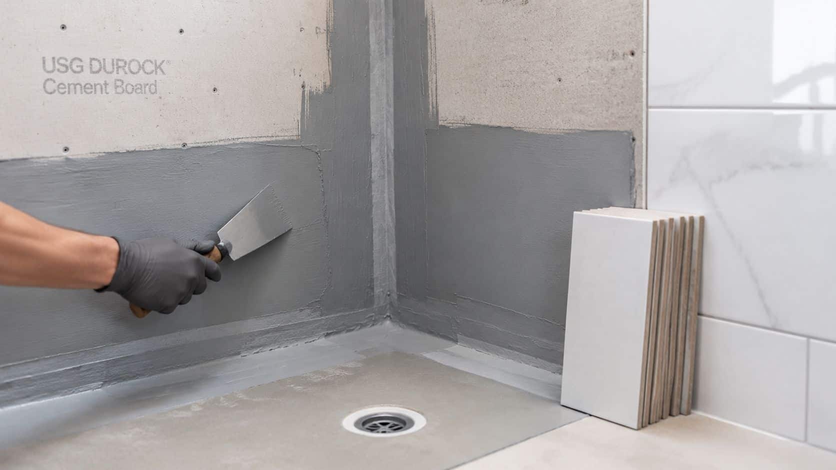 A gloved hand applies waterproof sealant to a cement board shower wall during a bathroom renovation.